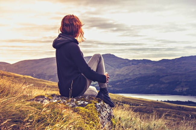 Mujer sentada en la ladera de una montaña contemplando el paisaje de las tierras altas escocesas